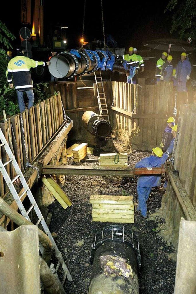 Construction workers using a crane to lower a large pipe into a shored trench at night