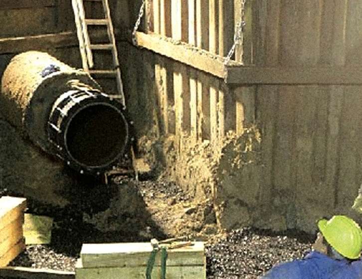 Construction worker in a yellow hard hat inside a shored trench beside a large underground pipe