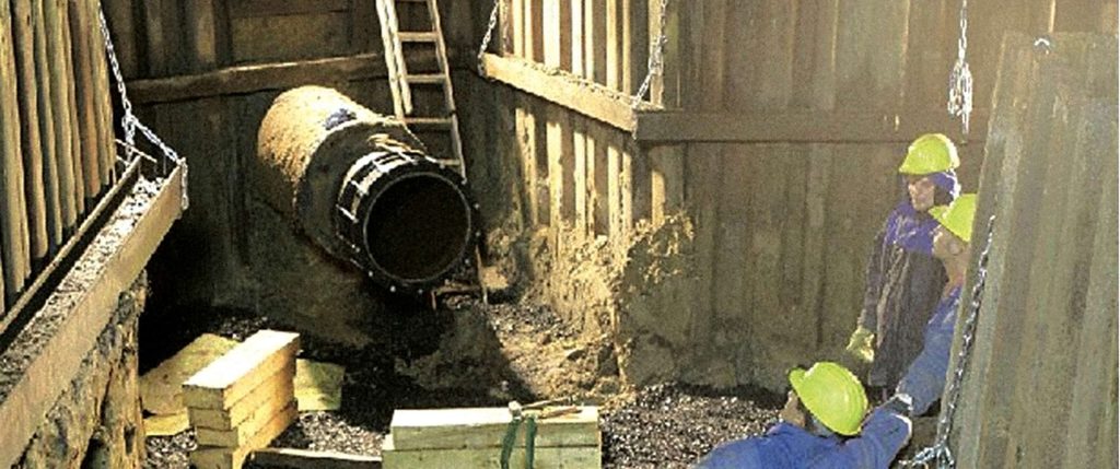 Construction workers wearing yellow hard hats working inside a timber-shored trench near a large underground pipe