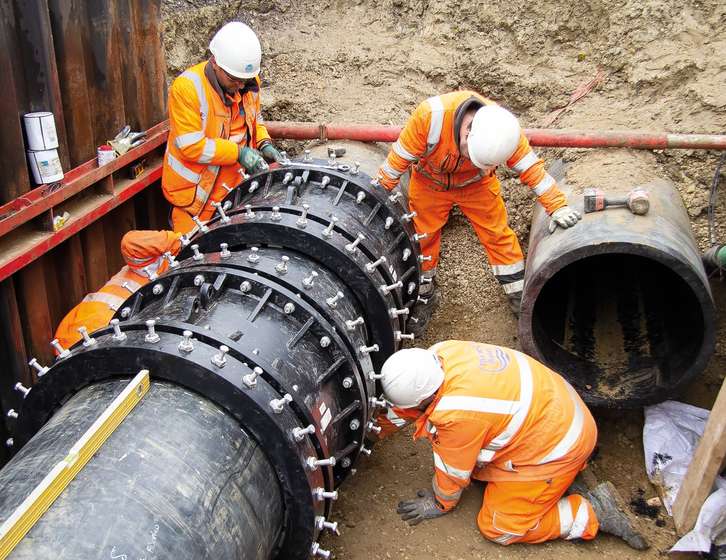 Three construction workers in orange hi-vis gear fitting large bolted couplings to underground pipes in a trench