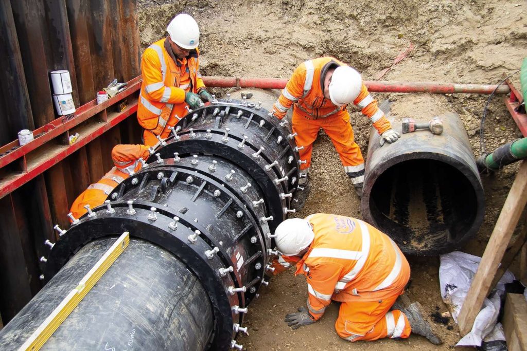 Construction workers in orange hi-vis clothing installing large bolted pipe couplings in a trench