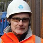 Construction worker wearing a white hard hat, tinted safety glasses, and a high-visibility jacket