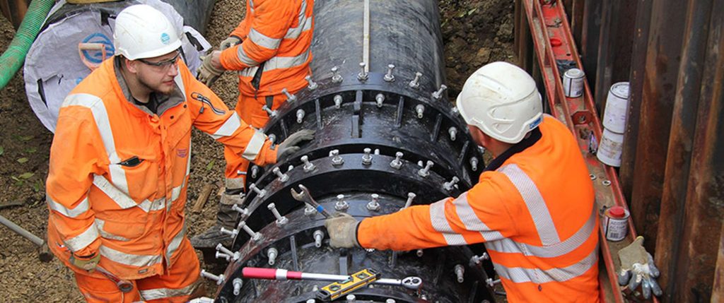 Construction workers in high-visibility clothing and hard hats installing a large bolted pipe joint in a trench