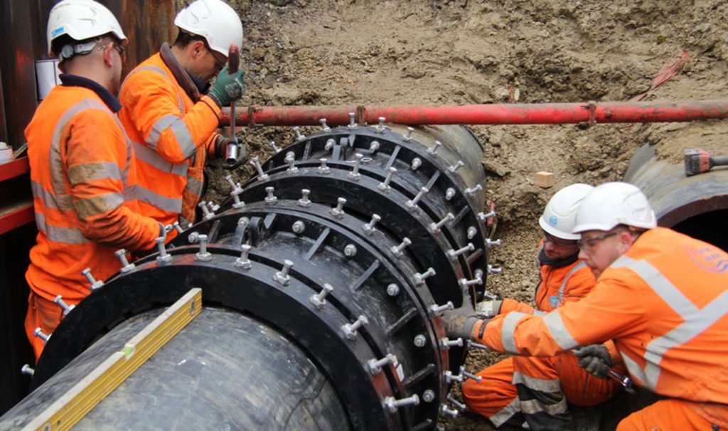 Construction workers in orange hi-vis gear and hard hats securing bolts on a large pipe joint in a trench
