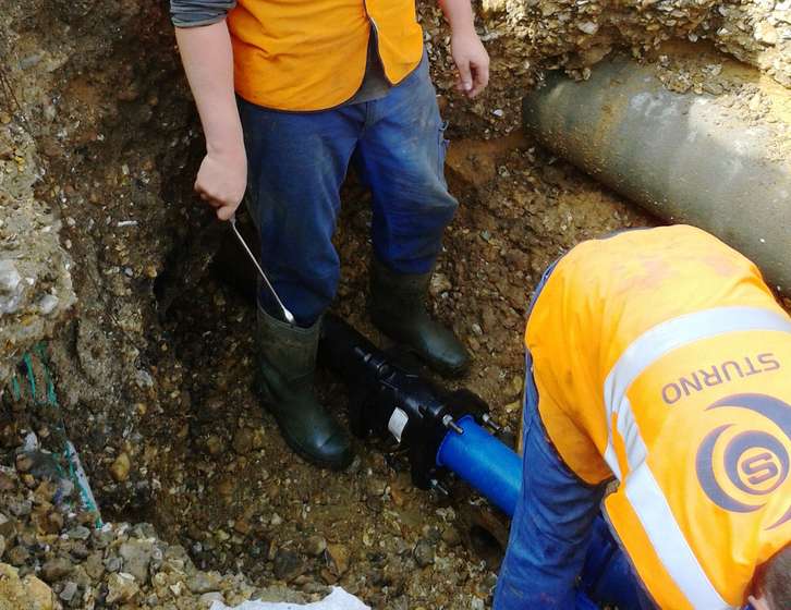 Two utility workers in high-visibility vests installing a blue pipe in an underground trench