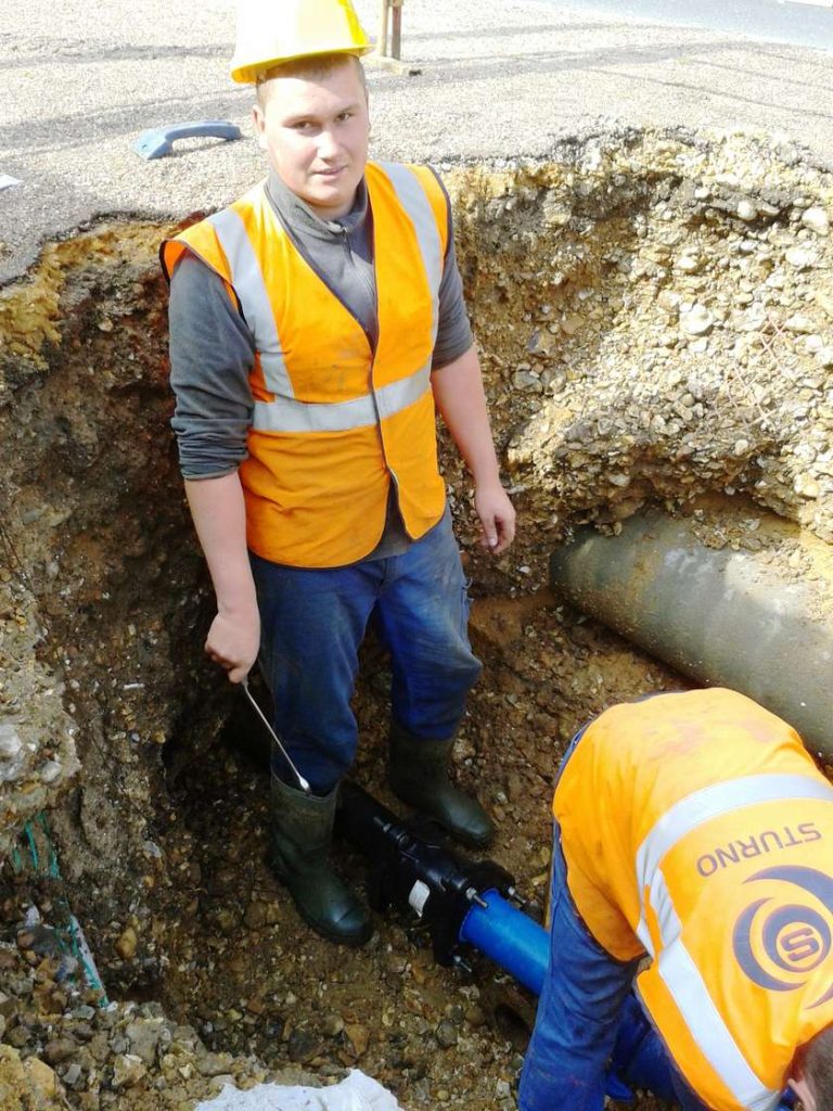 A construction worker standing in an excavated trench alongside a colleague repairing an underground pipe