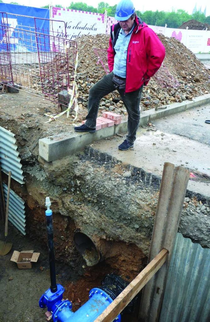 A construction worker in a hard hat and red jacket looking down into an excavated trench with exposed blue pipes