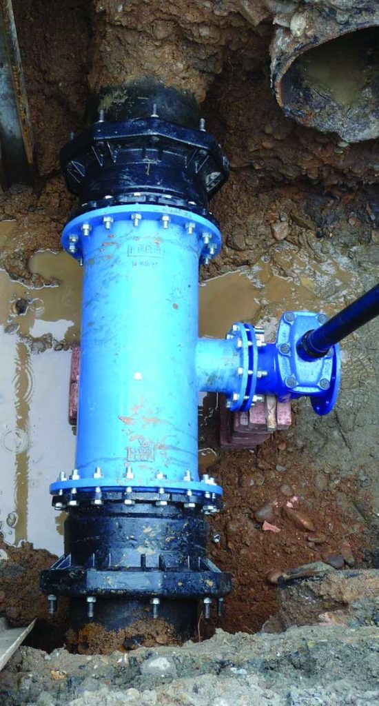 Large blue T-junction pipe fitted with bolted flanges inside a muddy excavation trench