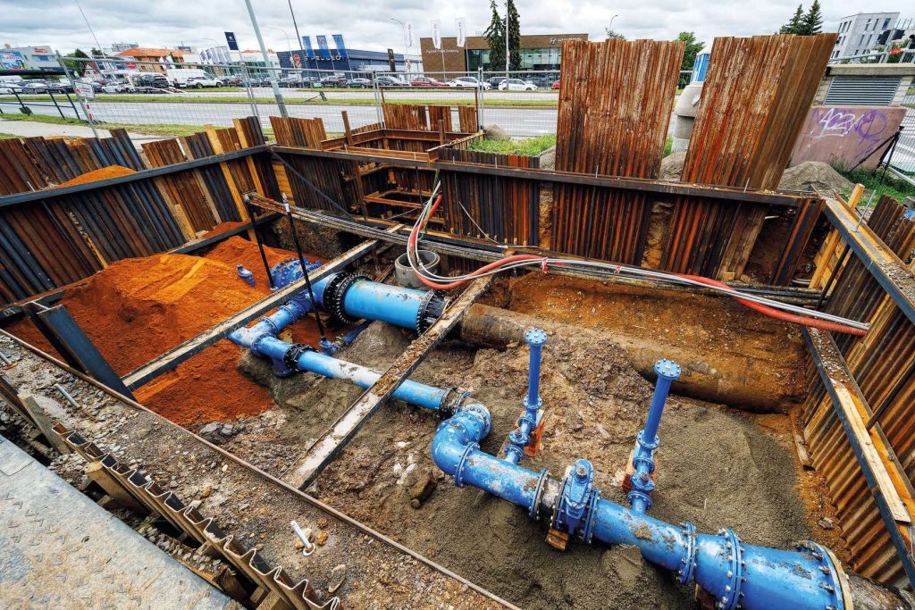 Excavated construction pit lined with steel sheet piling, exposing installed blue water pipes