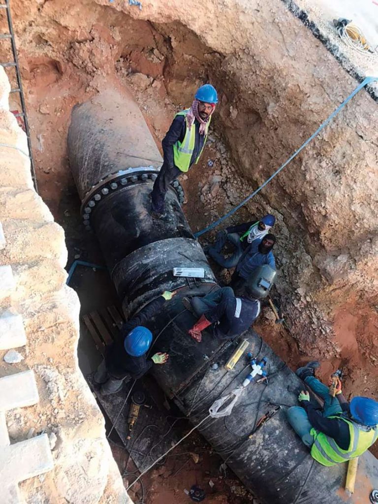 Construction workers in hard hats work on a large underground pipeline inside a deep trench