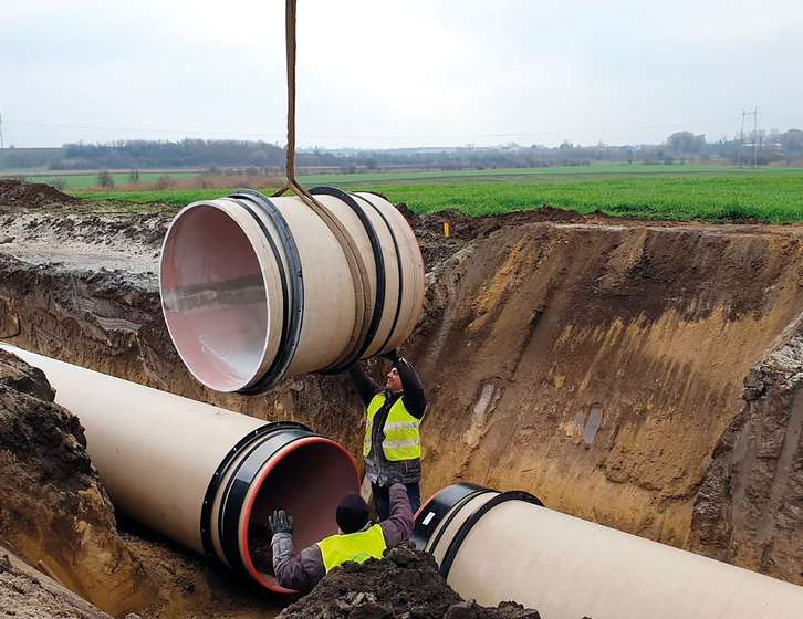 Two construction workers guiding a large pipe section being lowered by crane into a trench