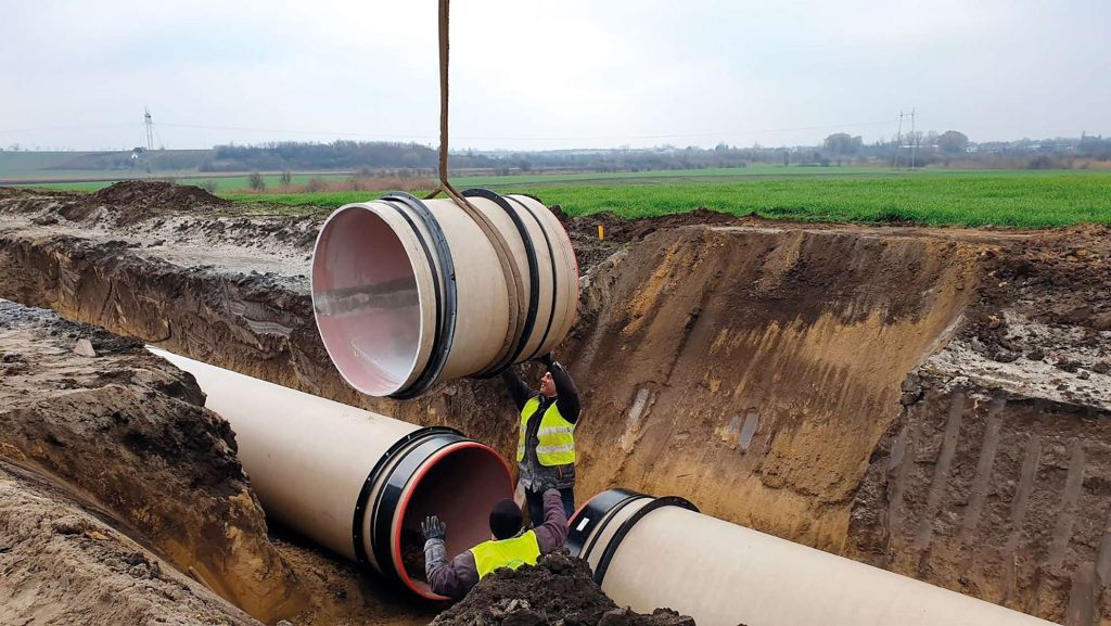 Construction workers in high-visibility jackets guiding a large pipe section into a trench