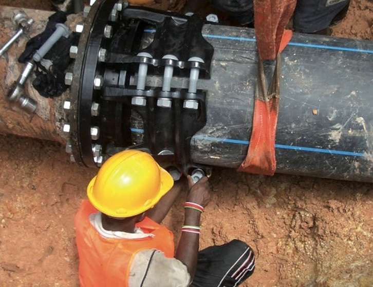 A worker in a yellow hard hat tightening bolts on a heavy-duty mechanical pipe coupling in a trench