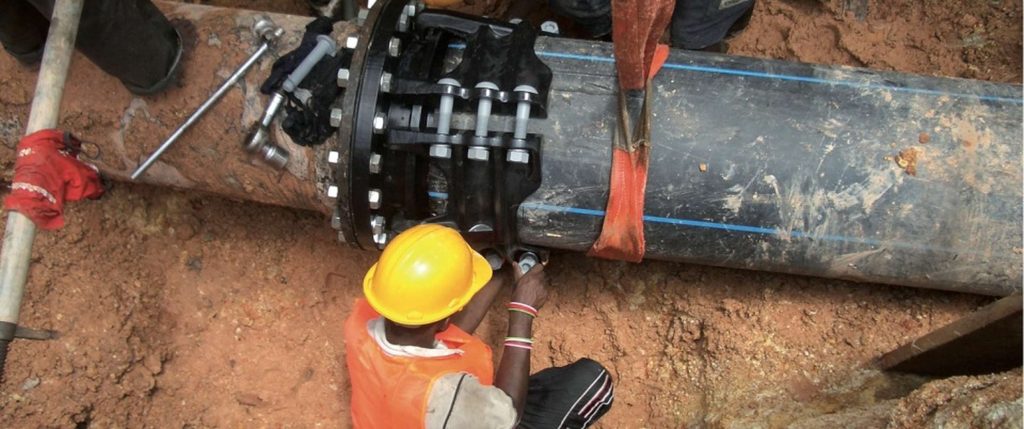 A worker in a hard hat and high-visibility vest installs a large mechanical coupling onto a pipe in a trench