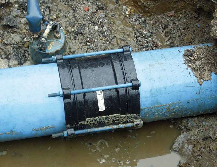 A black mechanical coupling clamp joining a large blue water pipe in a muddy trench