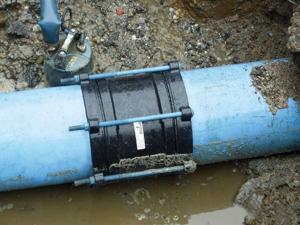 Black pipe repair coupling clamped onto a blue water pipe in a muddy trench
