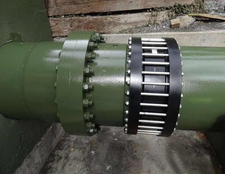 Large green industrial pipe showing a bolted flange connection and a black flexible expansion joint with silver tie rods
