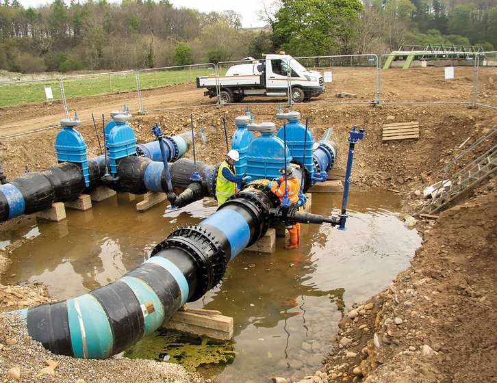 Two workers in hi-vis jackets installing large blue valves on water pipes inside a flooded trench