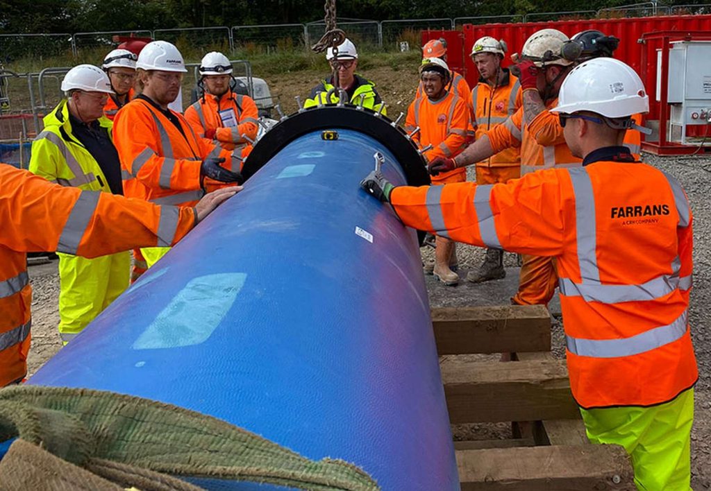 Construction workers wearing hi-vis jackets and hard hats installing a large blue pipe on site