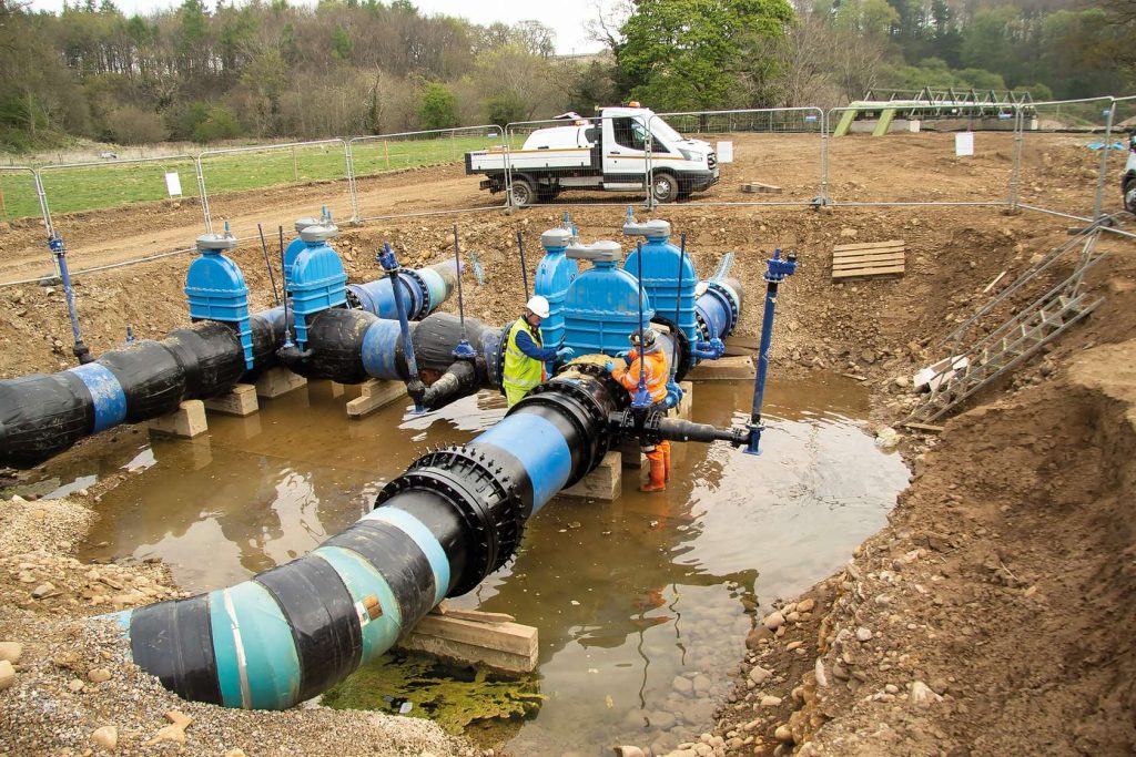 Two workers in hi-vis gear working on large blue valves and black water pipes inside a flooded excavation pit