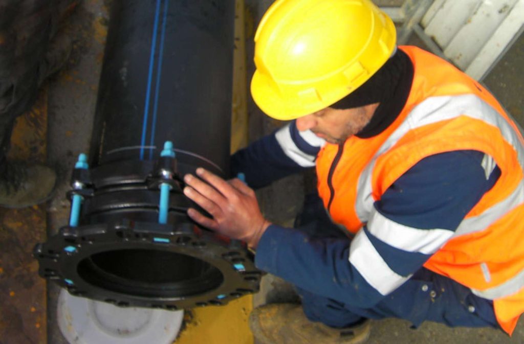 A worker wearing a yellow hard hat and high-visibility vest fitting a flange coupling onto a large black pipe