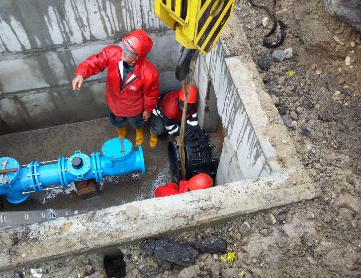 Workers in red waterproofs guiding a pipe component into an underground trench using a crane hook