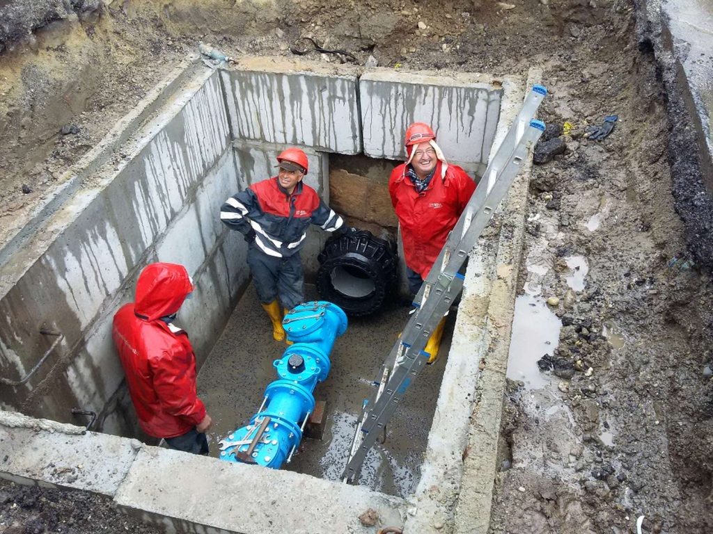 Three utility workers in red jackets inside a concrete trench installing blue pipes and valves