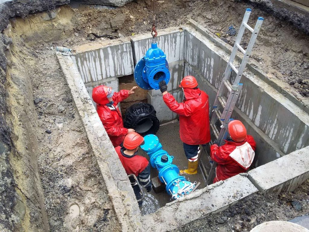 Four construction workers in red jackets installing a large blue water valve inside a concrete pit