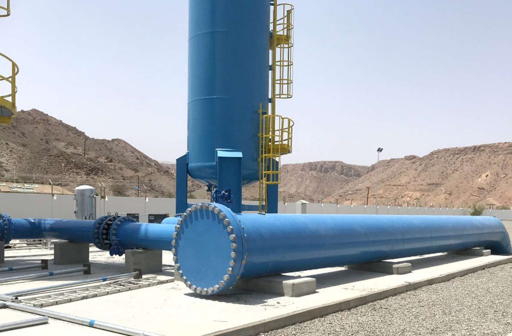 Large blue industrial pipes and vertical tank with a yellow safety ladder, set against desert mountains