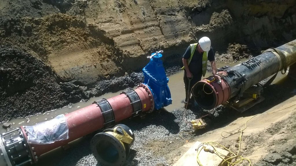 Worker inspecting large water pipes and a blue gate valve in an excavated trench