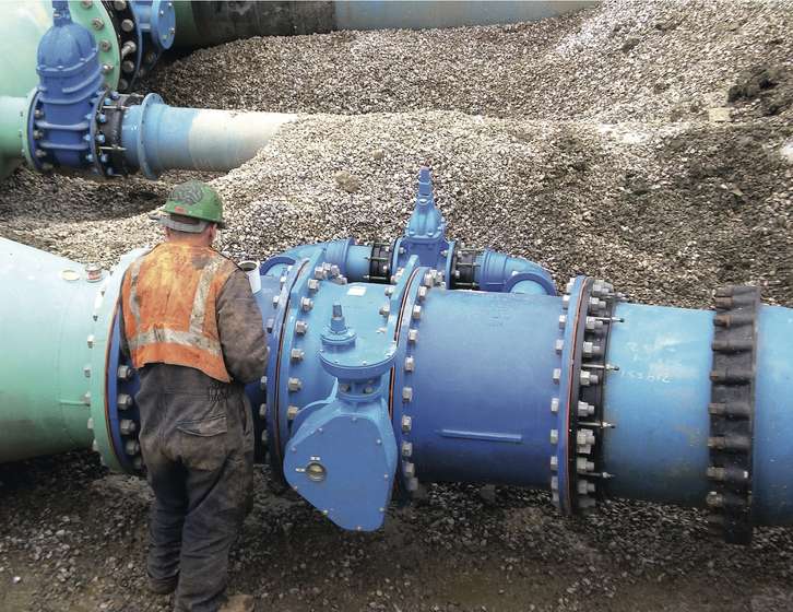 Construction worker standing beside large blue industrial water pipes and valves in a gravel trench
