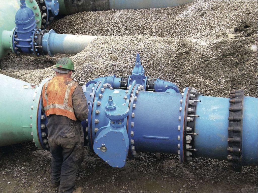 A construction worker inspecting large blue pipeline valves installed in a gravel trench