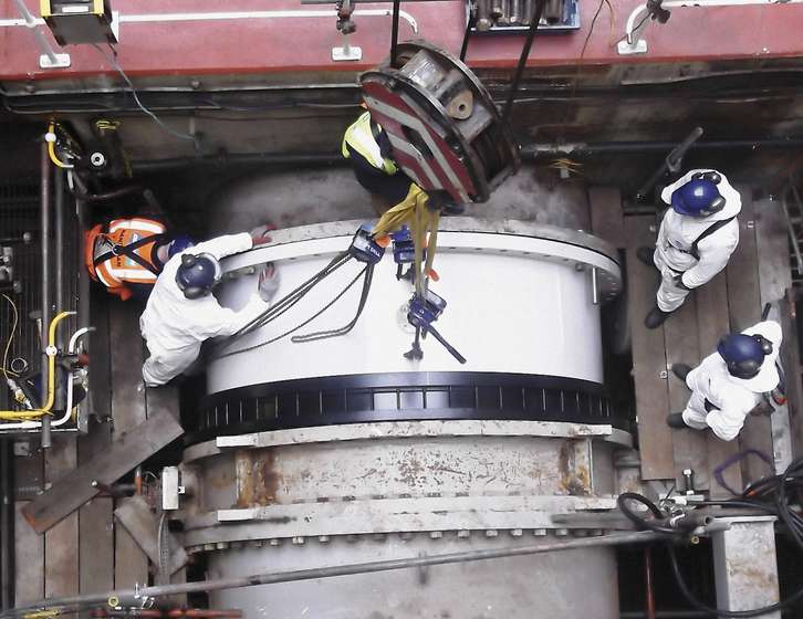 Overhead view of workers in white protective suits guiding a large cylindrical component into place with a crane hoist
