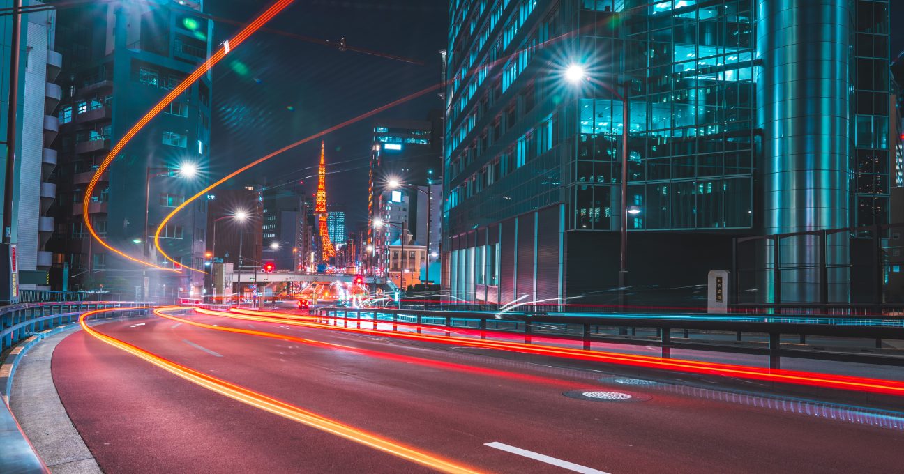 Long exposure of traffic light trails curving along a Tokyo street at night, with Tokyo Tower lit up in the background